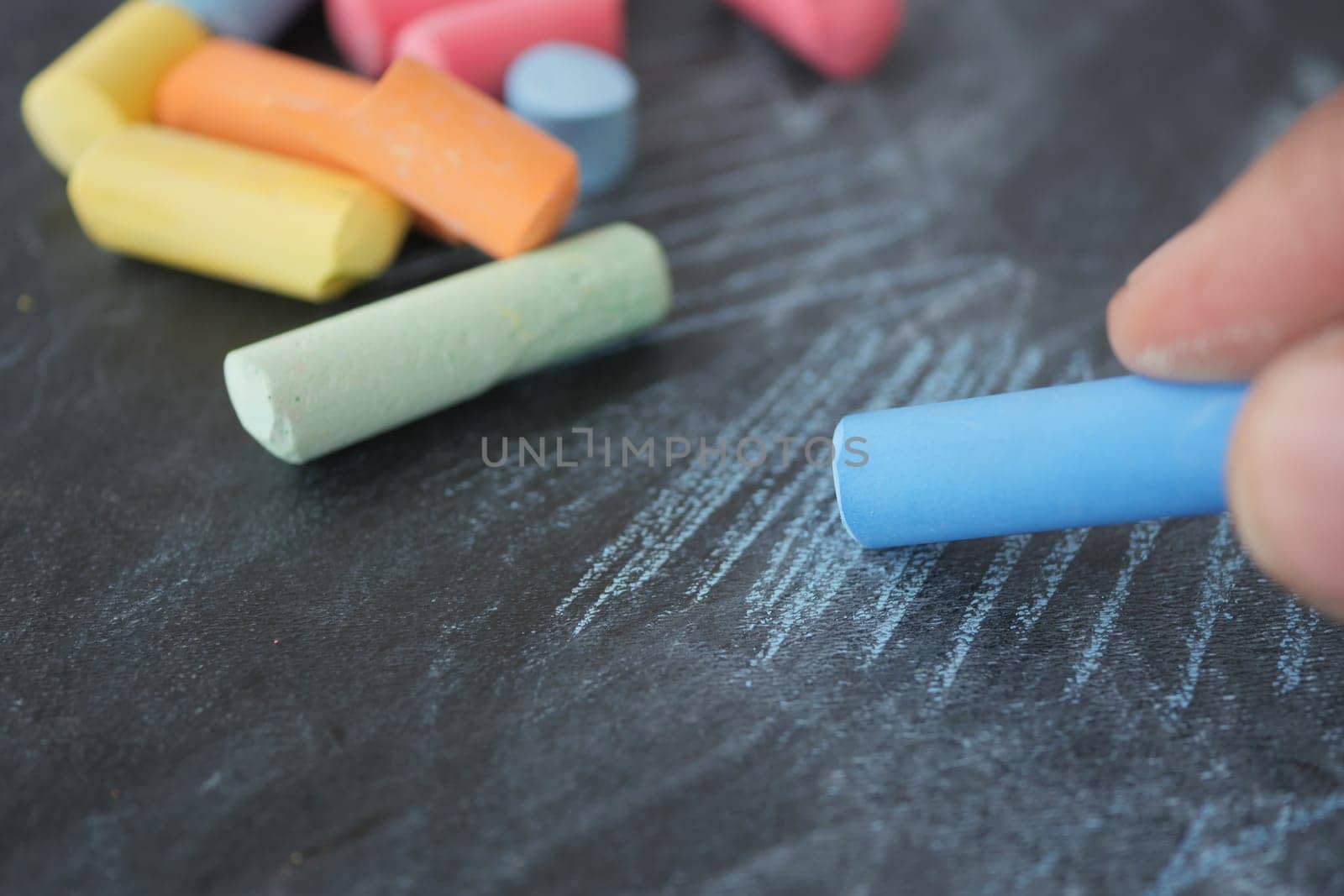 A hand draws with blue chalk on a dark surface, surrounded by colorful chalks.