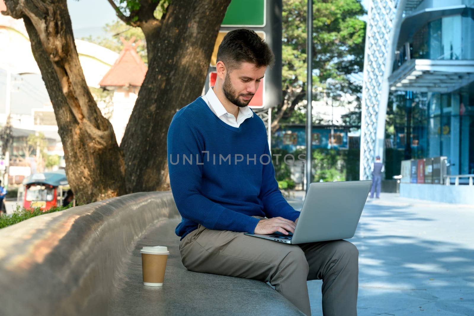 Caucasian male freelancer working on his laptop sitting on a curved outdoor bench.