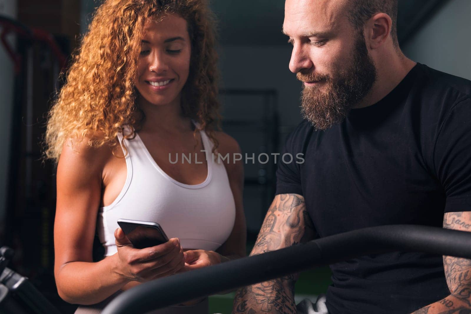 latin woman with curly hair in white top showing smartphone to man with tattoos in room, concept of technology of communication