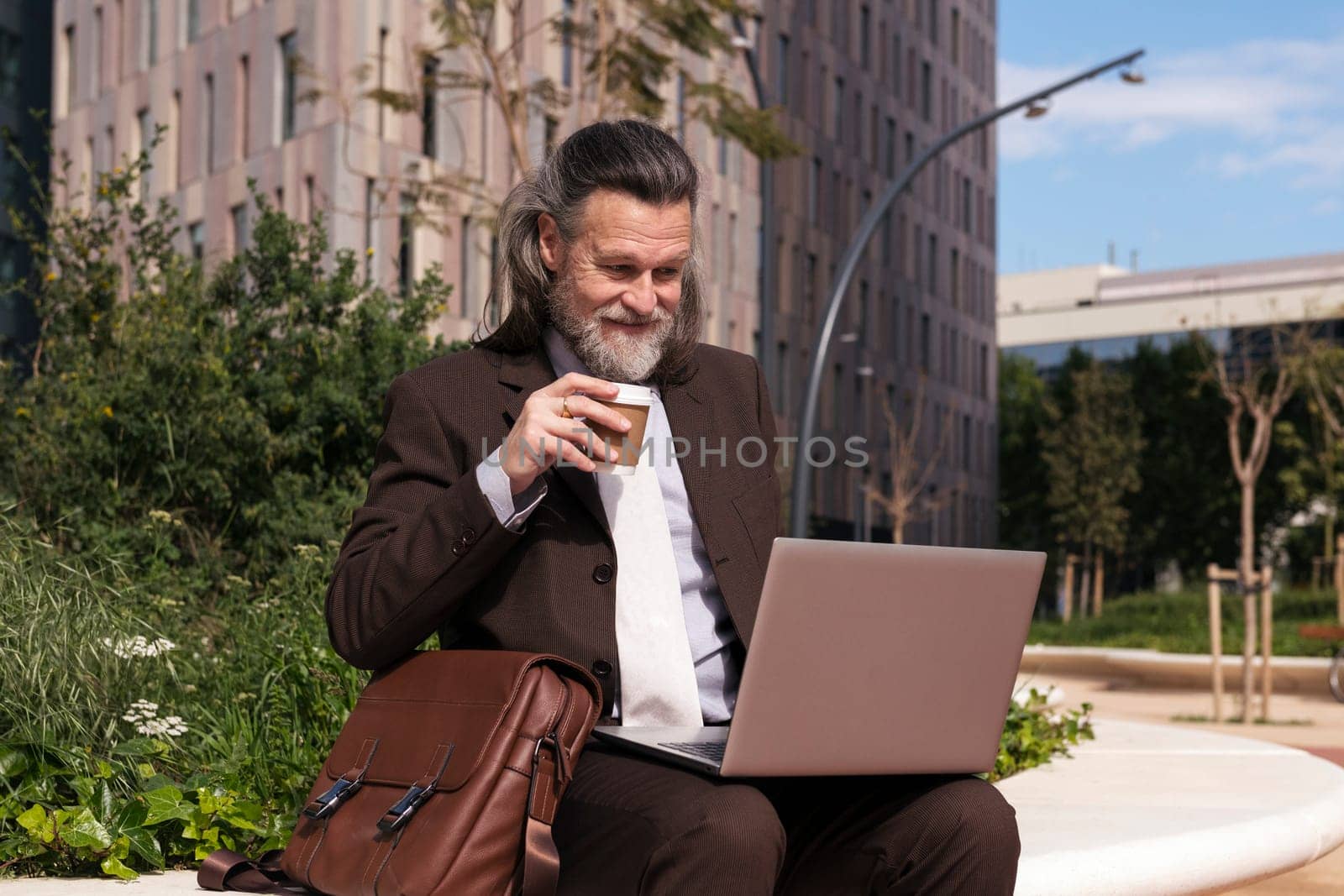 happy successful mature male in elegant clothes using laptop while drinking a cup of takeaway coffee sitting on urban square, concept of business and middle age man lifestyle