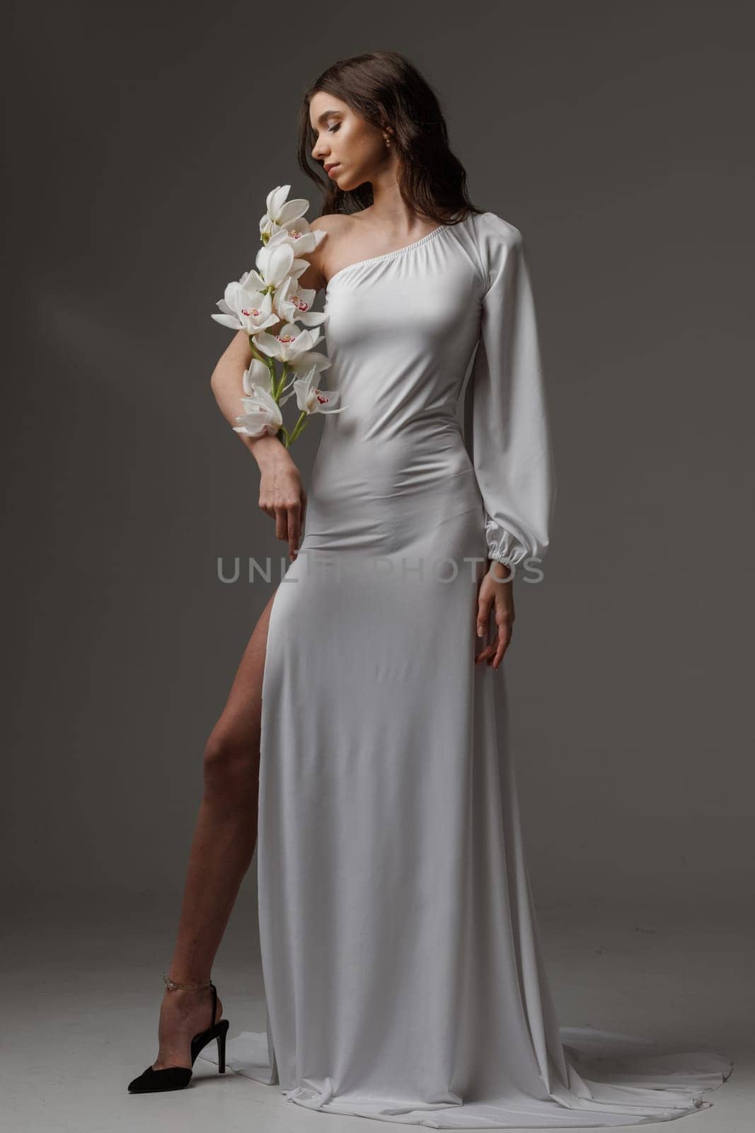 Elegant woman in a white one-shoulder dress holding white orchids, exuding grace, femininity, and sophistication in a minimalist studio setting.