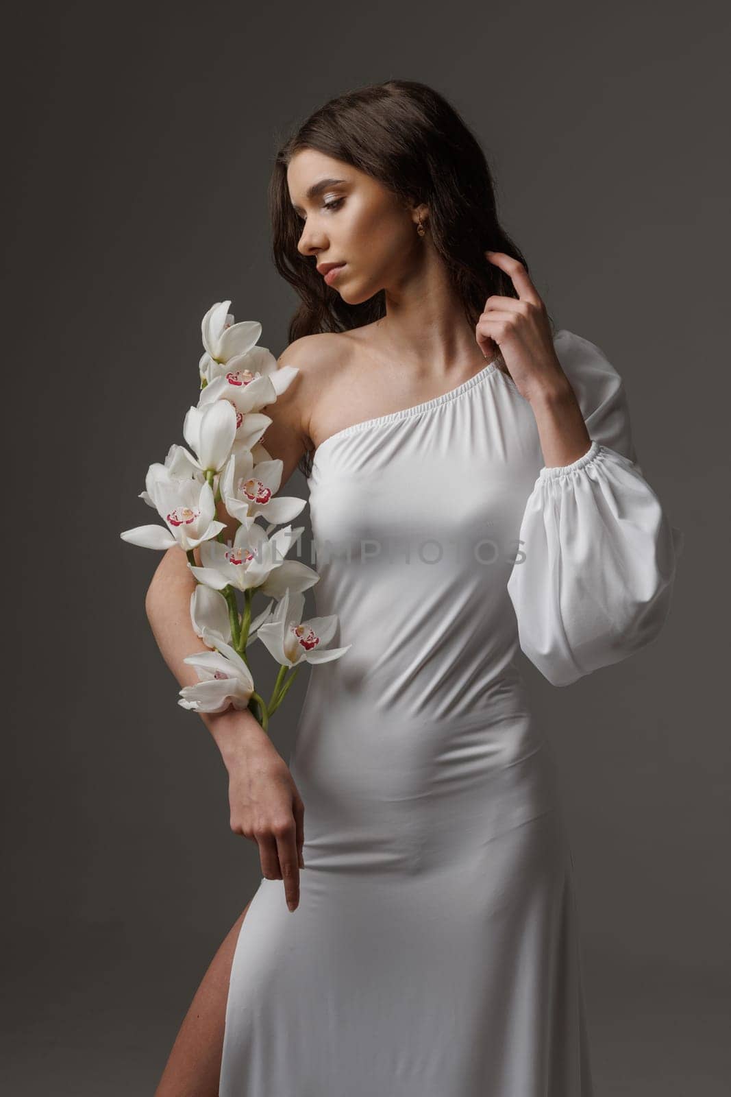 Elegant woman in a white one-shoulder dress holding white orchids, exuding grace, femininity, and sophistication in a minimalist studio setting.