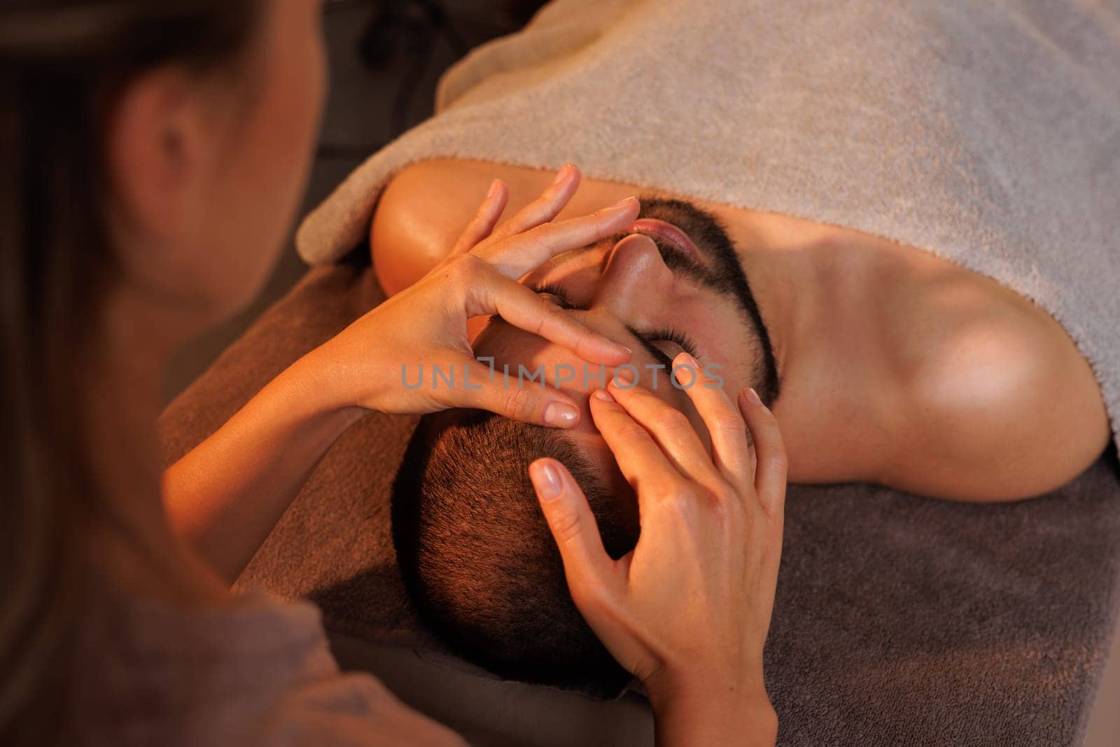 Relaxing facial massage for a bearded man in a spa, performed by a professional therapist. Warm lighting creates a soothing, wellness-focused atmosphere