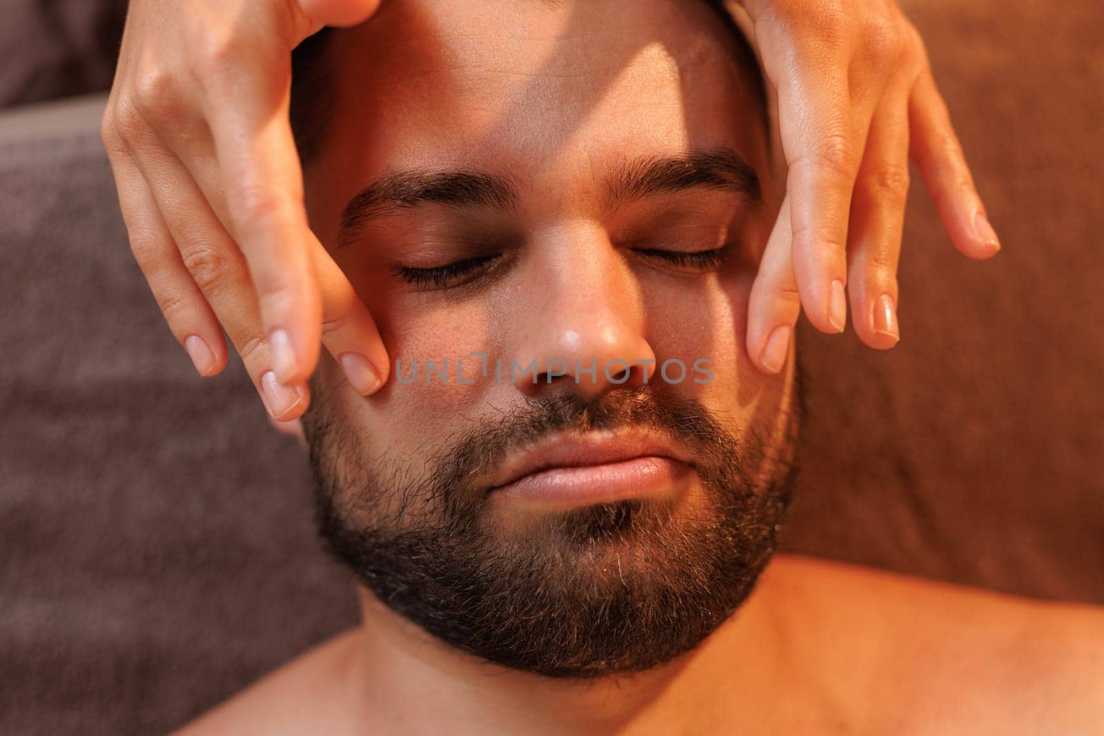 Close-up of a bearded man enjoying a relaxing facial massage at a spa. The therapists hands gently apply pressure, creating a soothing and rejuvenating self care experience,
