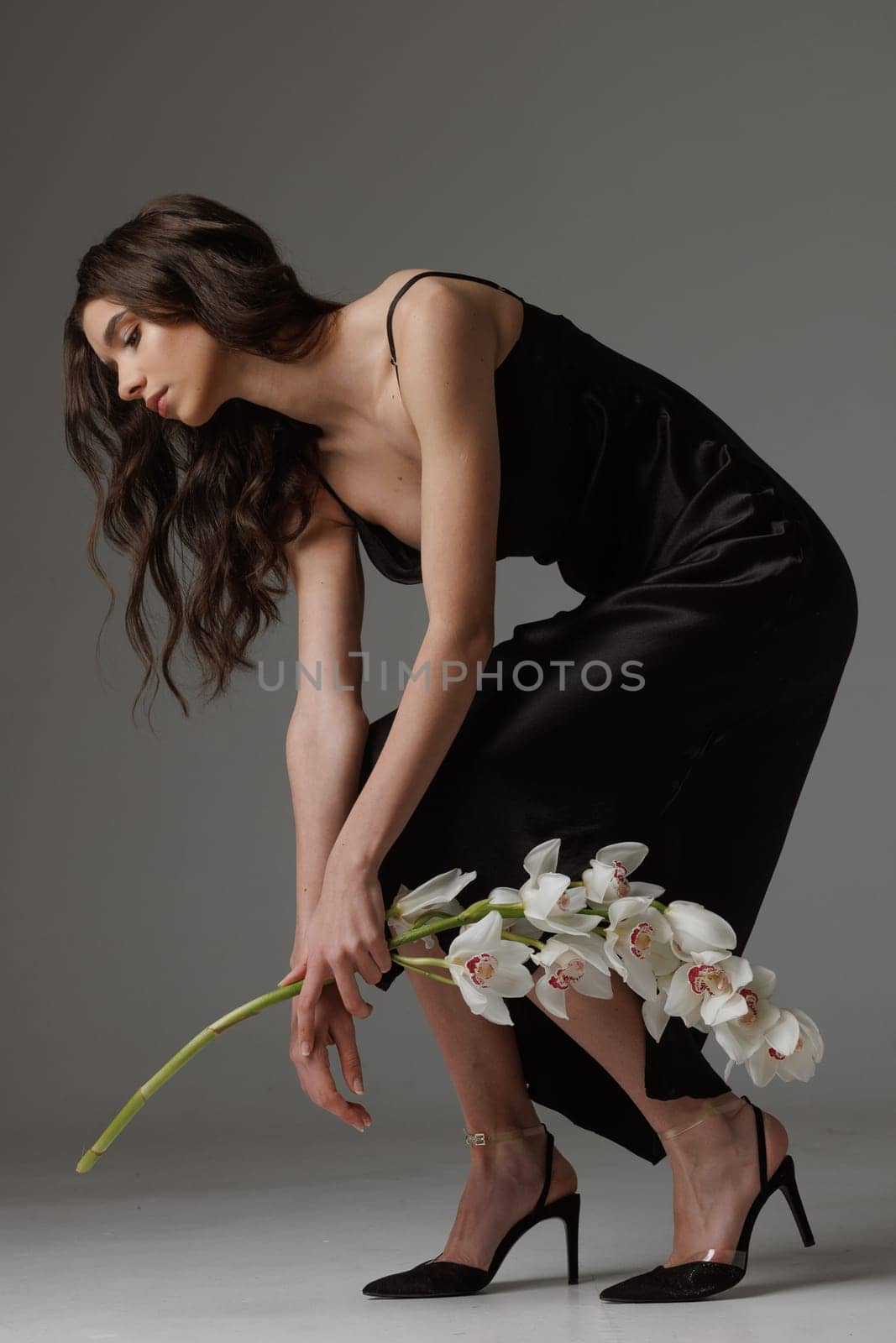 Elegant woman in a black satin dress bending gracefully while holding white orchids, showcasing beauty, fashion, and sophistication in a minimalist studio