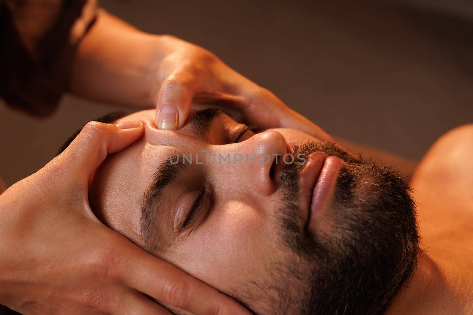 Close-up of a bearded man receiving a relaxing facial massage at a spa. Warm lighting enhances the peaceful and therapeutic atmosphere of this self-care and wellness treatment