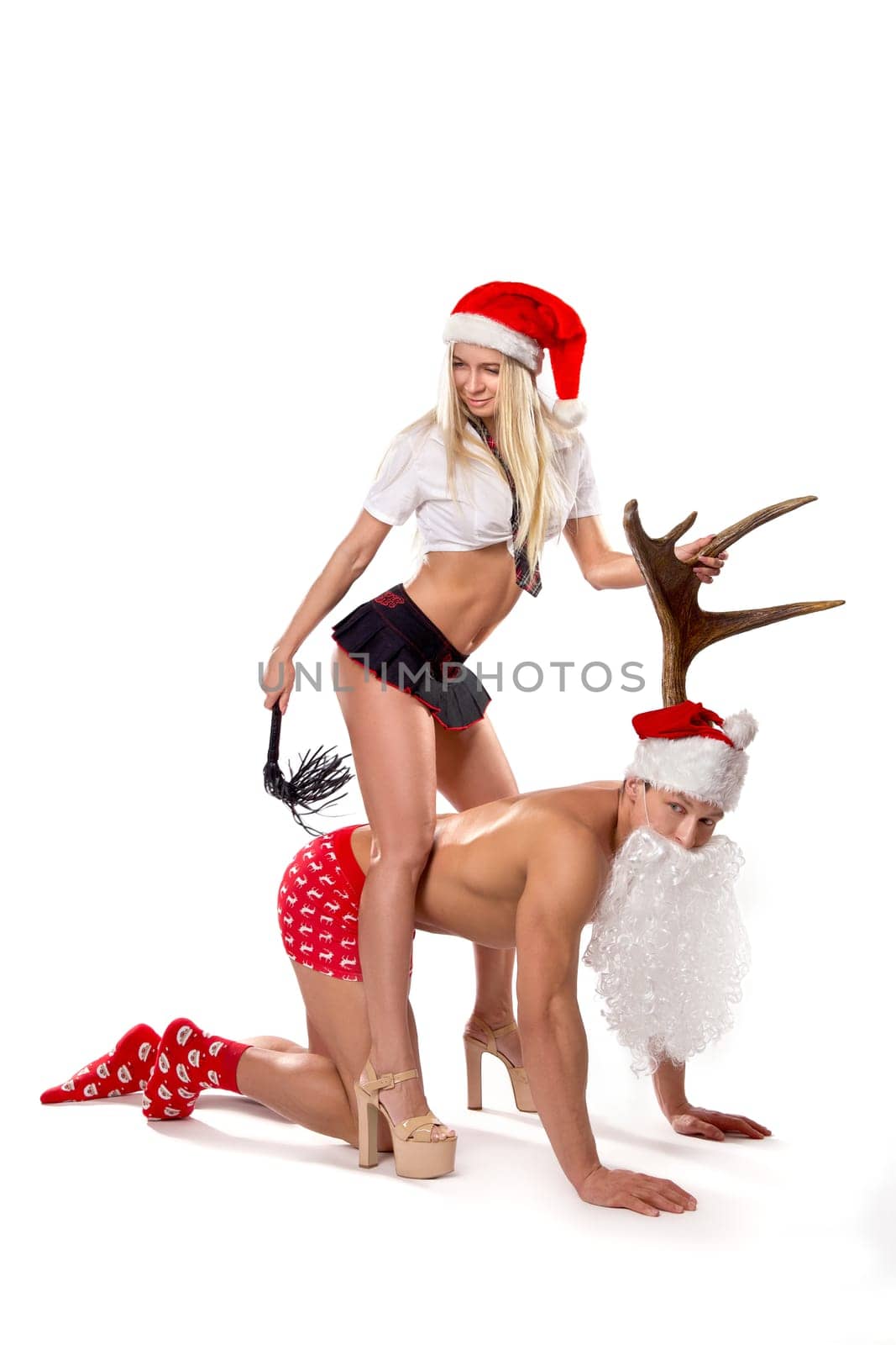 Full body of confident young blond female in sexy lingerie and high heels sitting on back of subordinate kneeling lover in Santa hat and leash
