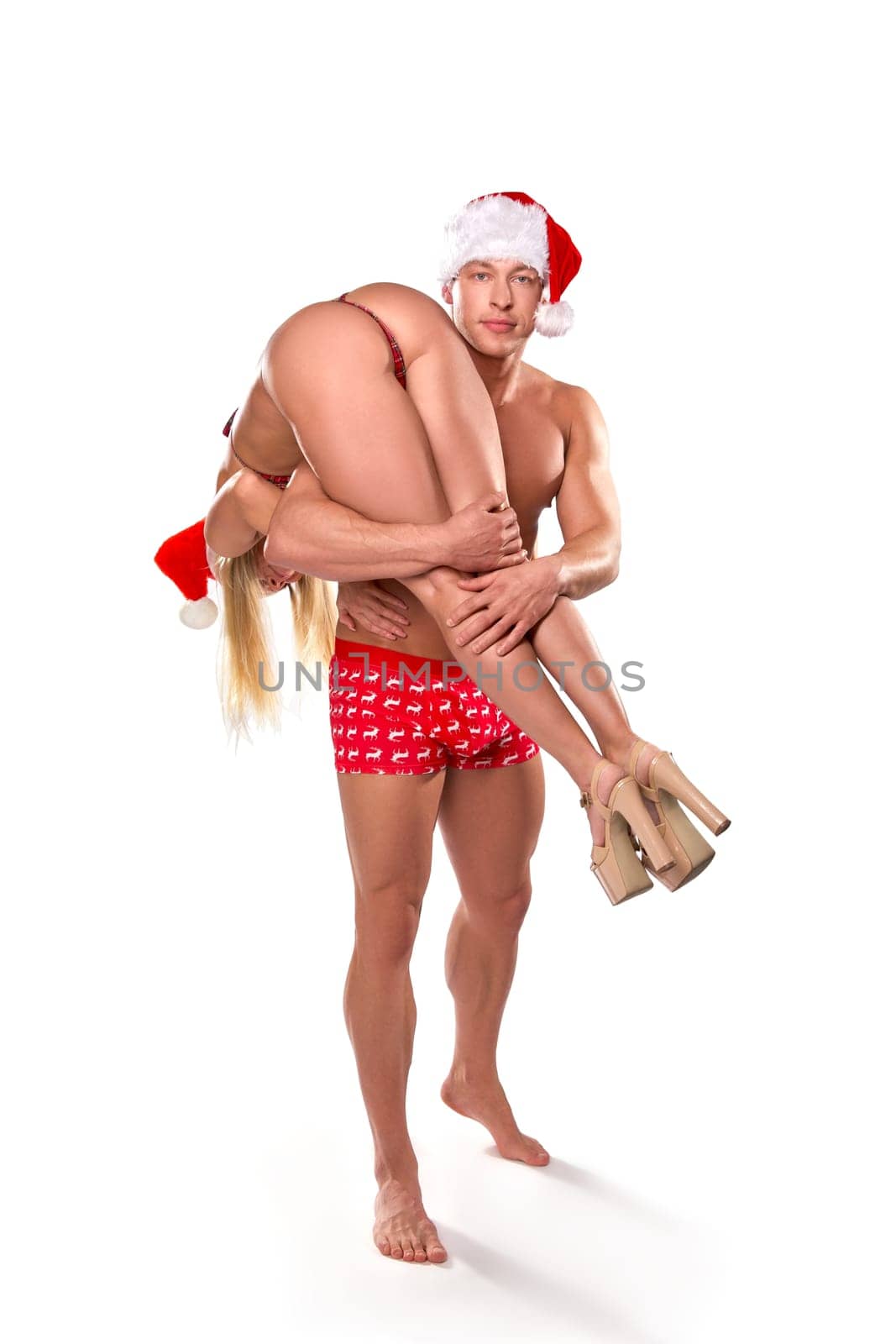 Full body of strong athletic handsome man in red underpants and Santa hat carrying sexy girlfriend with perfect body on white background