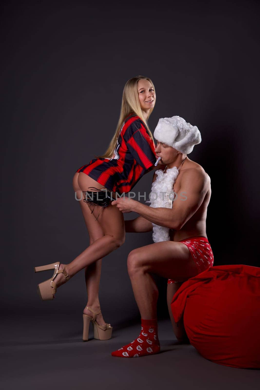 Side view of seductive blonde woman in football garment and high heels enjoying time with sexy boyfriend in Santa Claus russian style - bdsm theme