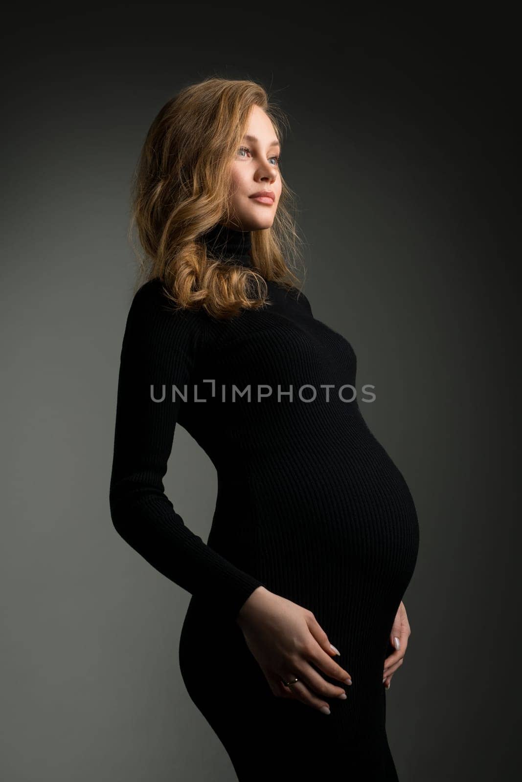 Side view of tranquil pregnant female wearing long black dress and touching tummy while standing on dark background in studio