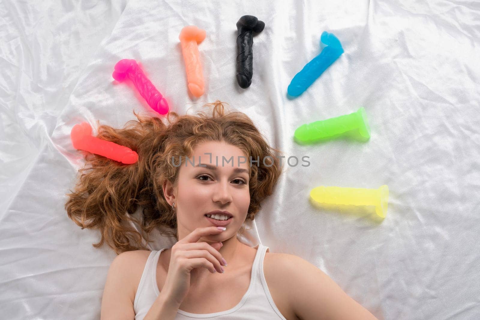 Top view of cheerful female relaxing on soft bed with colorful rubber dildos and looking at camera