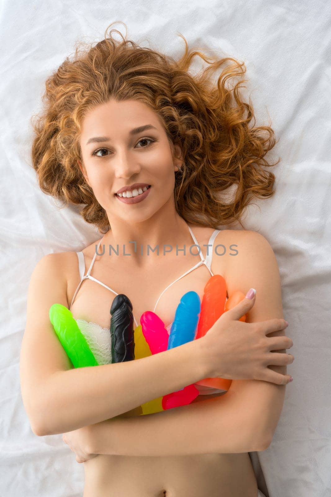 Top view of cheerful female relaxing on soft bed with colorful rubber dildos and looking at camera