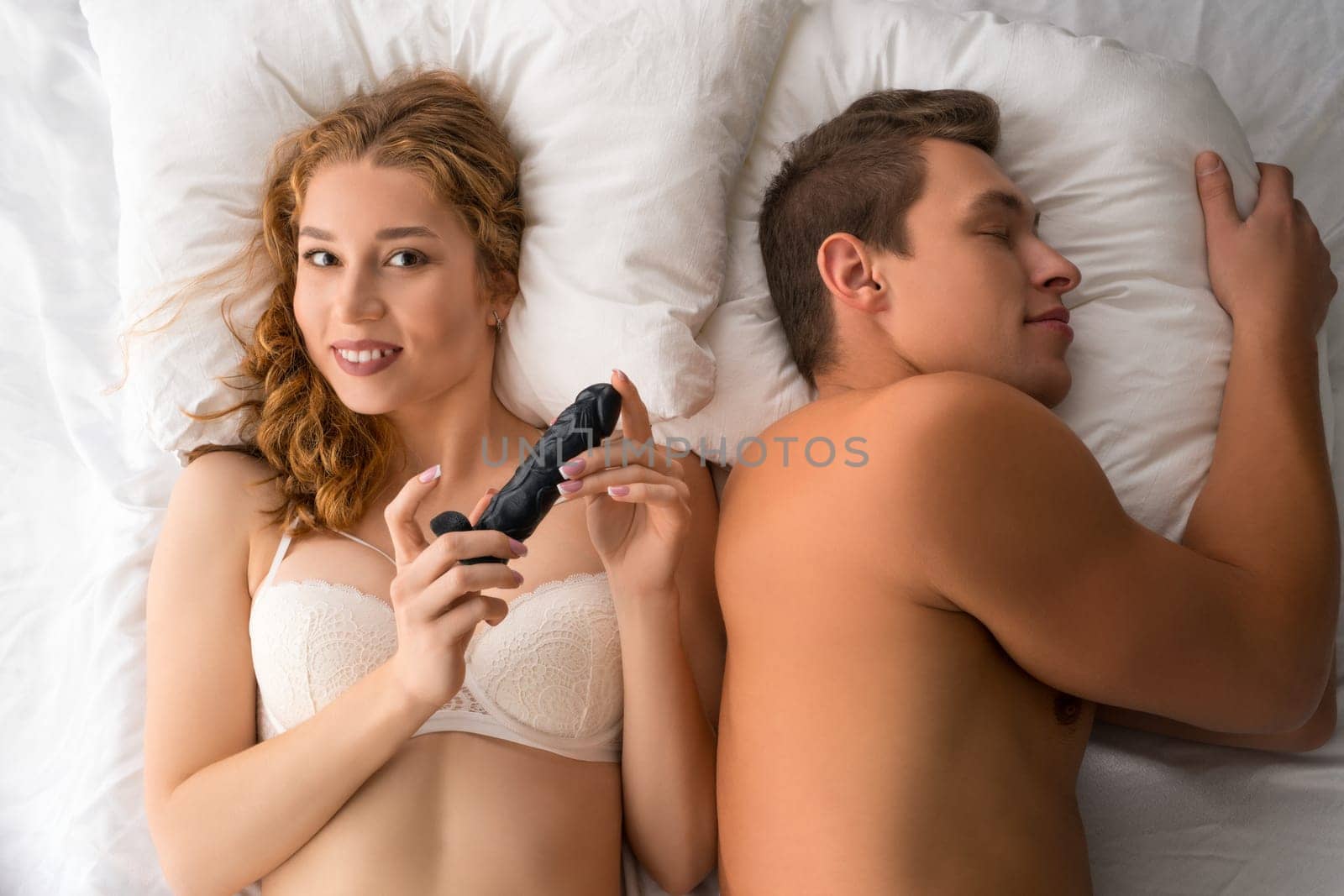 Top view of playful female in underwear lying with sexual toy on bed next to sleeping boyfriend