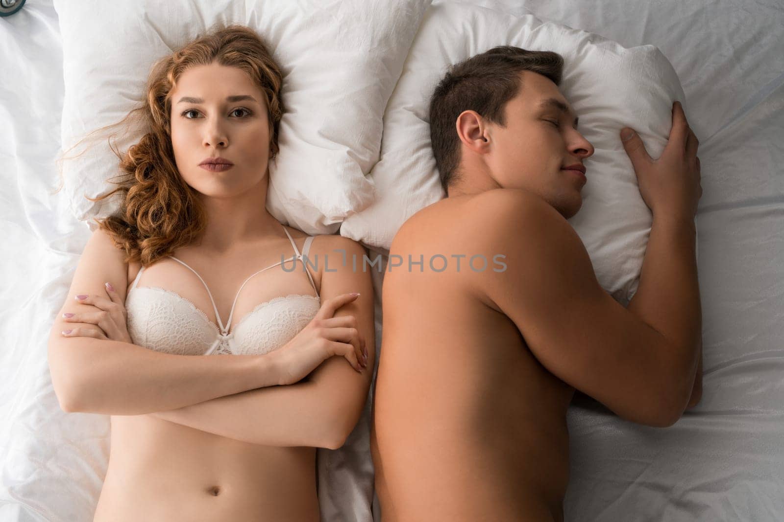 Top view of discontent female in underwear lying with crossed arms on bed next to sleeping boyfriend