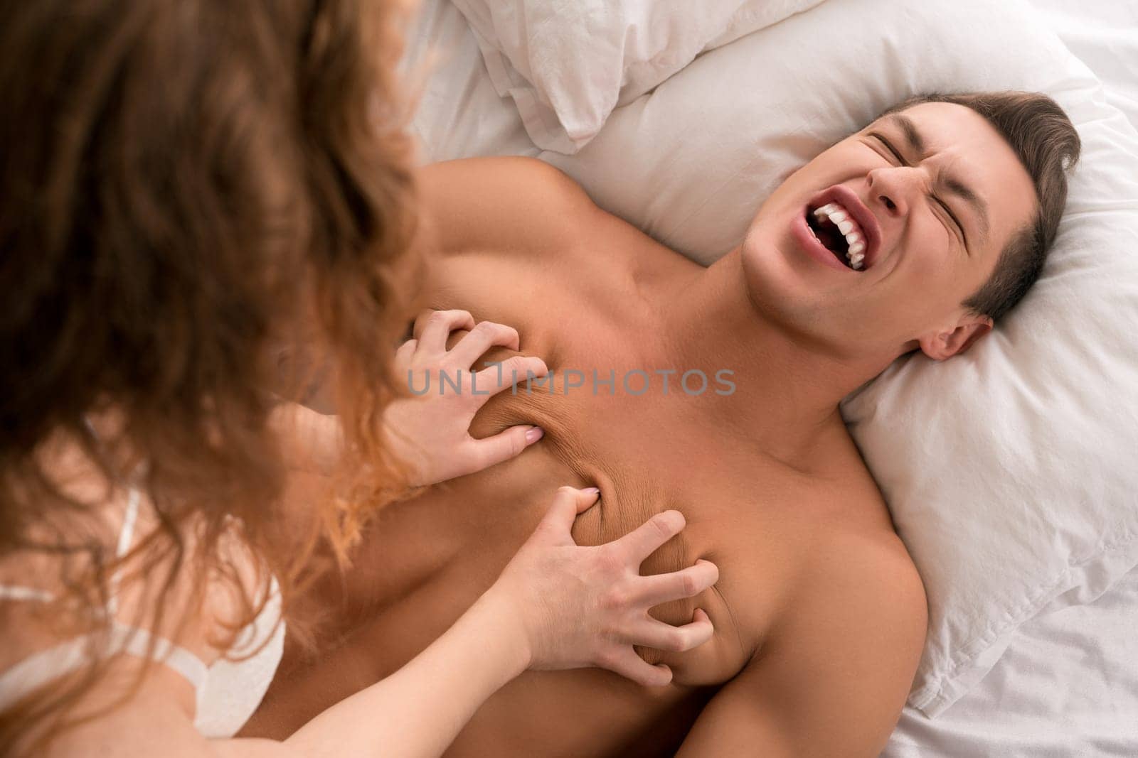 High angle of anonymous woman touching bare breast of man crying from pain sitting on bed and enjoying intimate moments