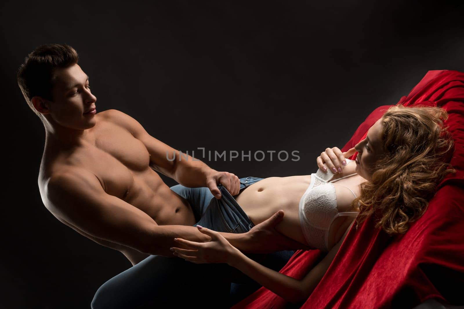 Muscular shirtless man taking off pants from slim seductive woman while having sex on red sheets against black background