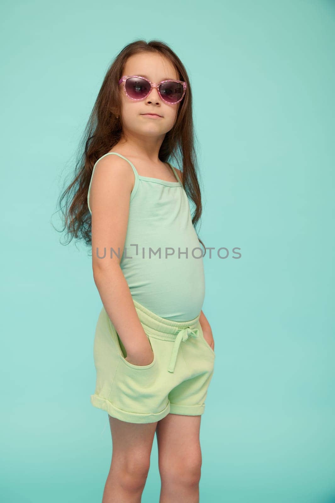 Vertical studio portrait of a Caucasian cute little child girl in stylish eyeglasses, dressed in summer wear, looking confidently at the camera, isolated over blue background. Beautiful kids. Children