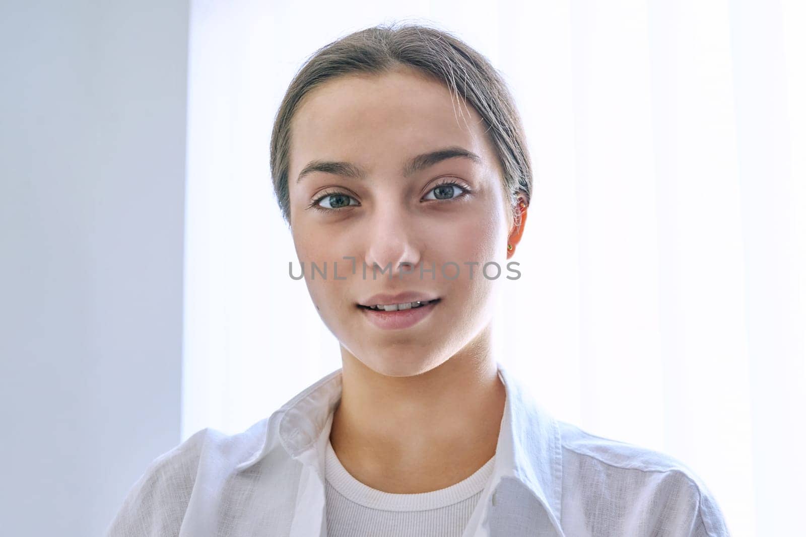 Headshot portrait of teen girl at home near window. Smiling female teenager 16, 17 years old looking at camera