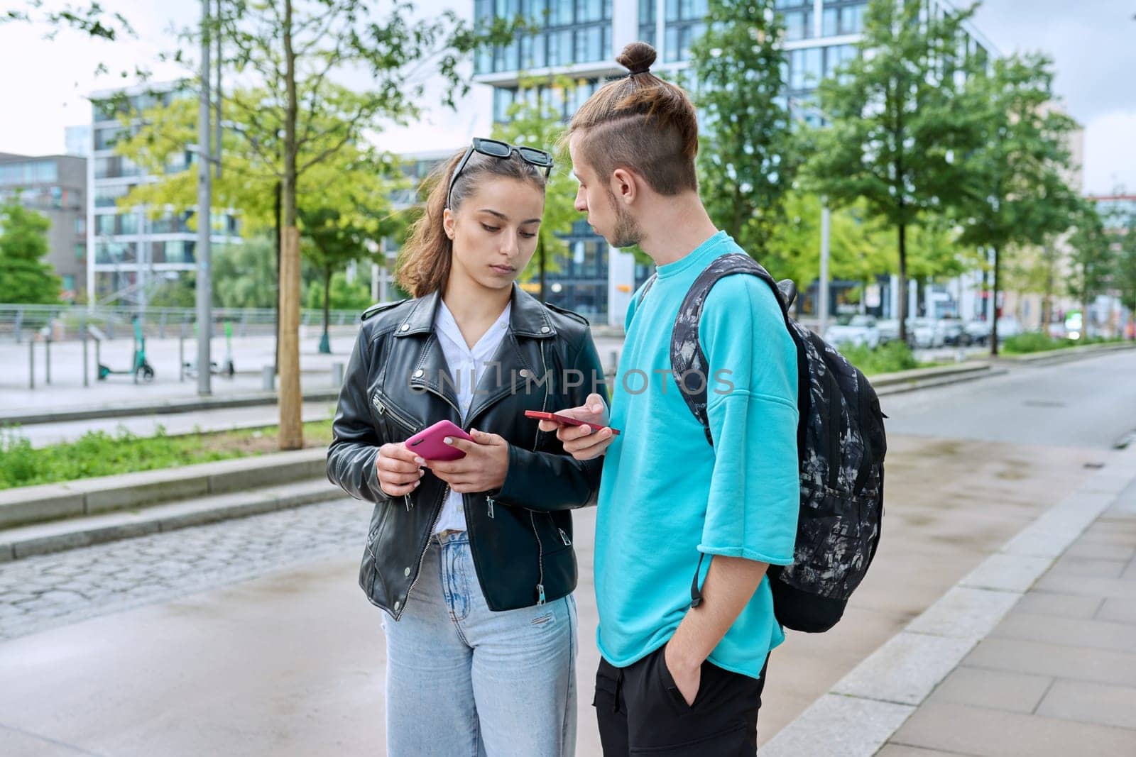 Teen friends guy and girl standing together holding smartphones looking at screen using mobile phones outdoor on city street. Internet digital technology applications for leisure study communication
