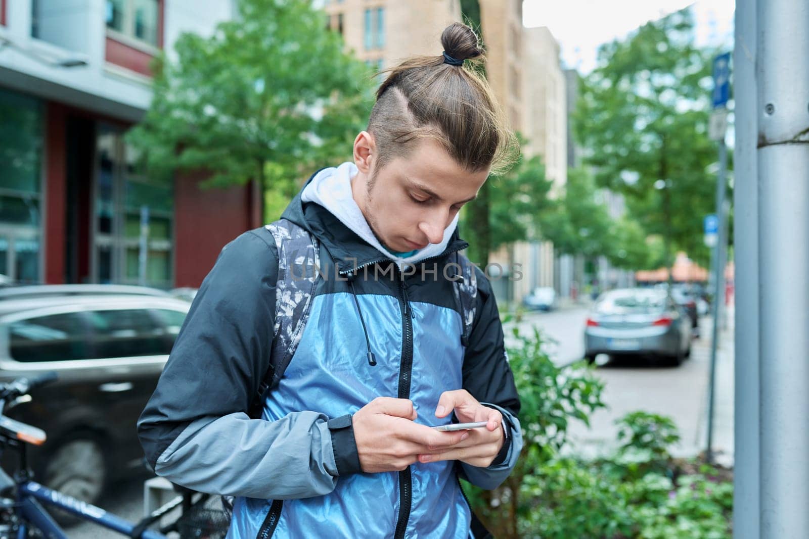 Fashionable young male with backpack using smartphone outdoors in city by VH-studio