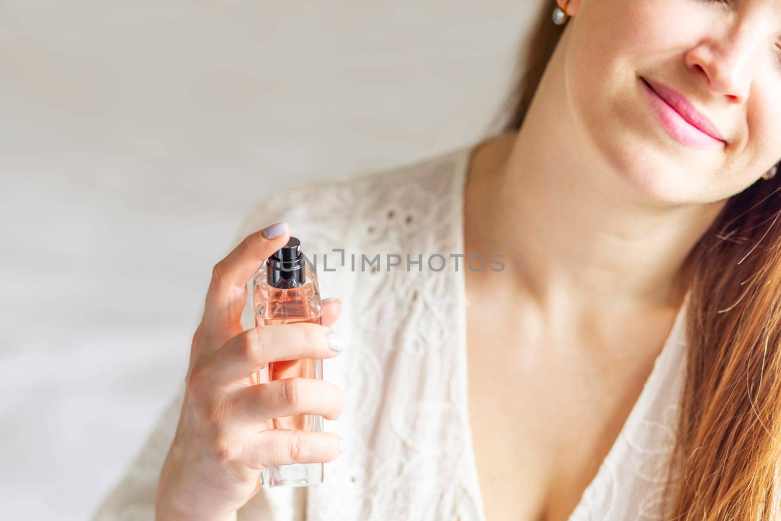 Beautiful young woman with bottle of perfume spraying it on her neck at home.