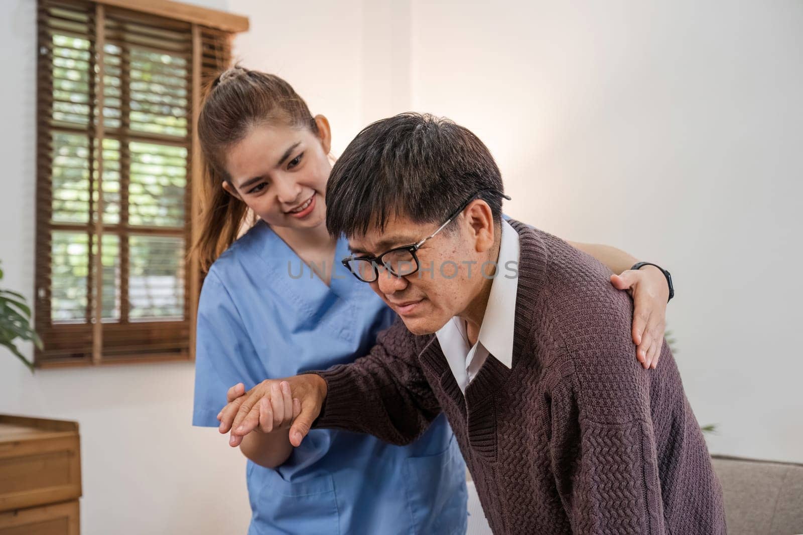 A caring young Asian nurse helps an elderly person walk in the house. An elderly disabled man tries to walk with the help of a female caregiver. by wichayada