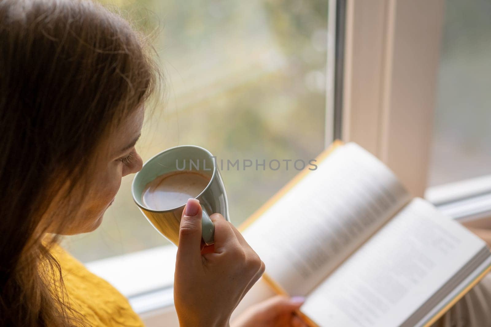 Young beautiful woman sitting by the window yellow knitted sweater read book, daily planner, notepad. Relax concept. Hold cappuccino glass of coffee with white foam. Text is out of focus