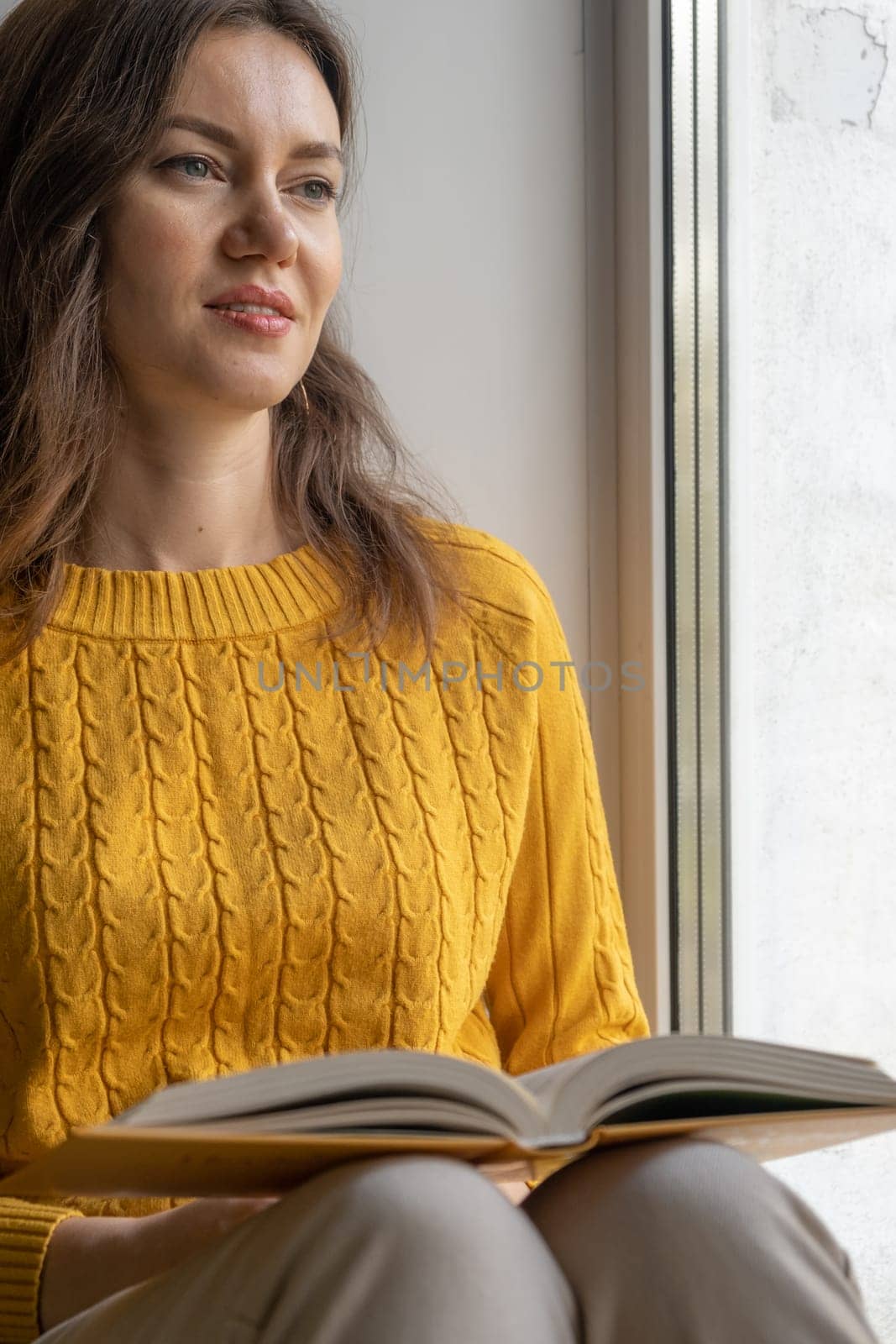 Young beautiful woman sitting by the window yellow knitted sweater read book, daily planner, notepad. Relax concept. Hold cappuccino glass of coffee with white foam. Text is out of focus