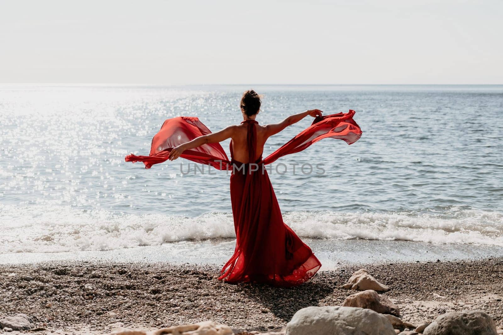 Side view a Young beautiful sensual woman in a red long dress posing on a rock high above the sea during sunrise. Girl on the nature on blue sky background. Fashion photo.