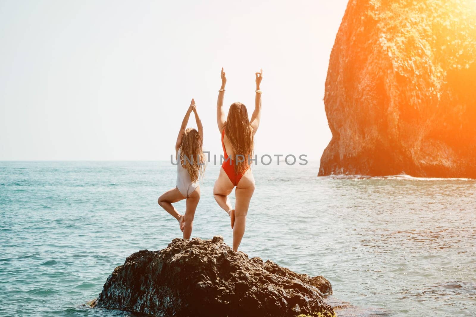 Woman and her daughter practicing balancing yoga pose on one leg up together on rock in the sea. Silhouette mother and daughter doing yoga at beach by panophotograph