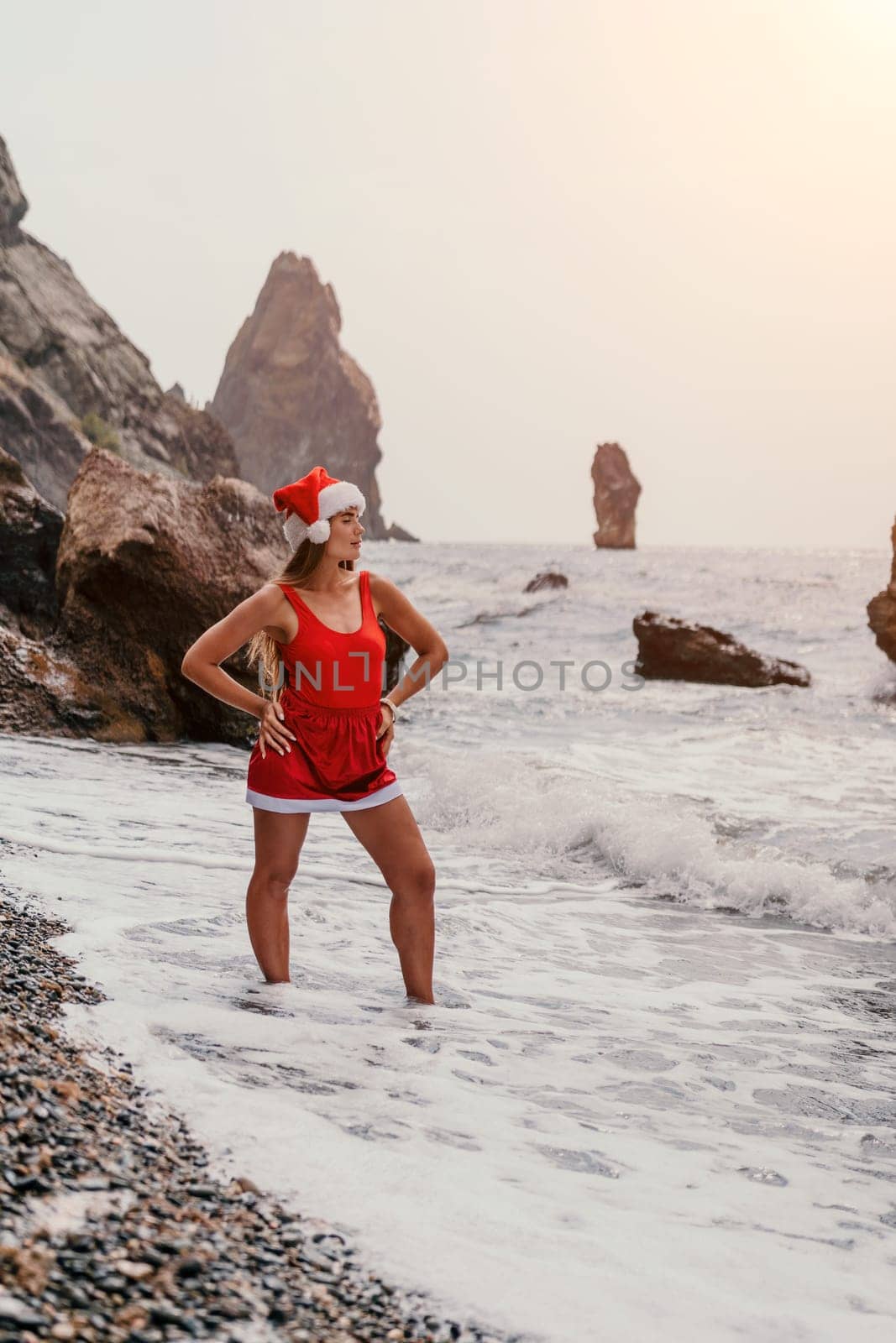 Woman summer travel sea. Happy tourist in red bikini and Santas hat enjoy taking picture outdoors for memories. Woman traveler posing on the beach surrounded by volcanic mountains, sharing travel joy by panophotograph