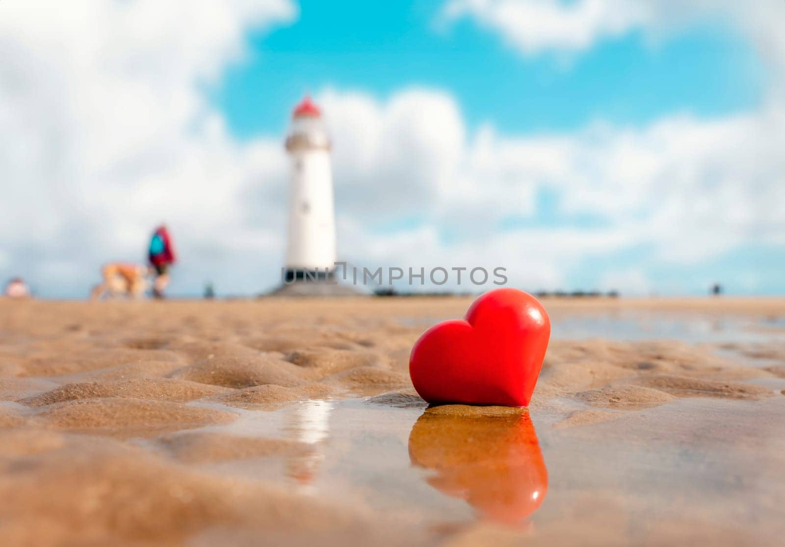 Big red heart in sand at coast against lighthouse. Fall in love with holiday at seaside concept