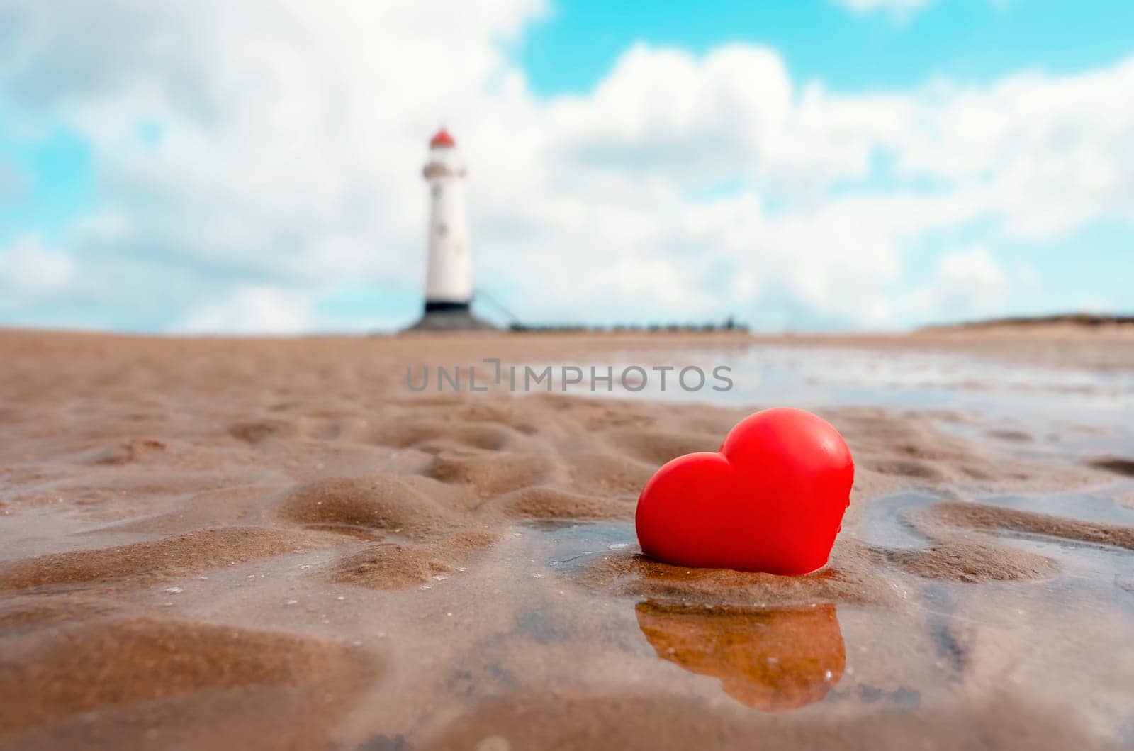 red heart in sand at coast against lighthouse. Fall in love with holiday at seaside concept by Iryna_Melnyk