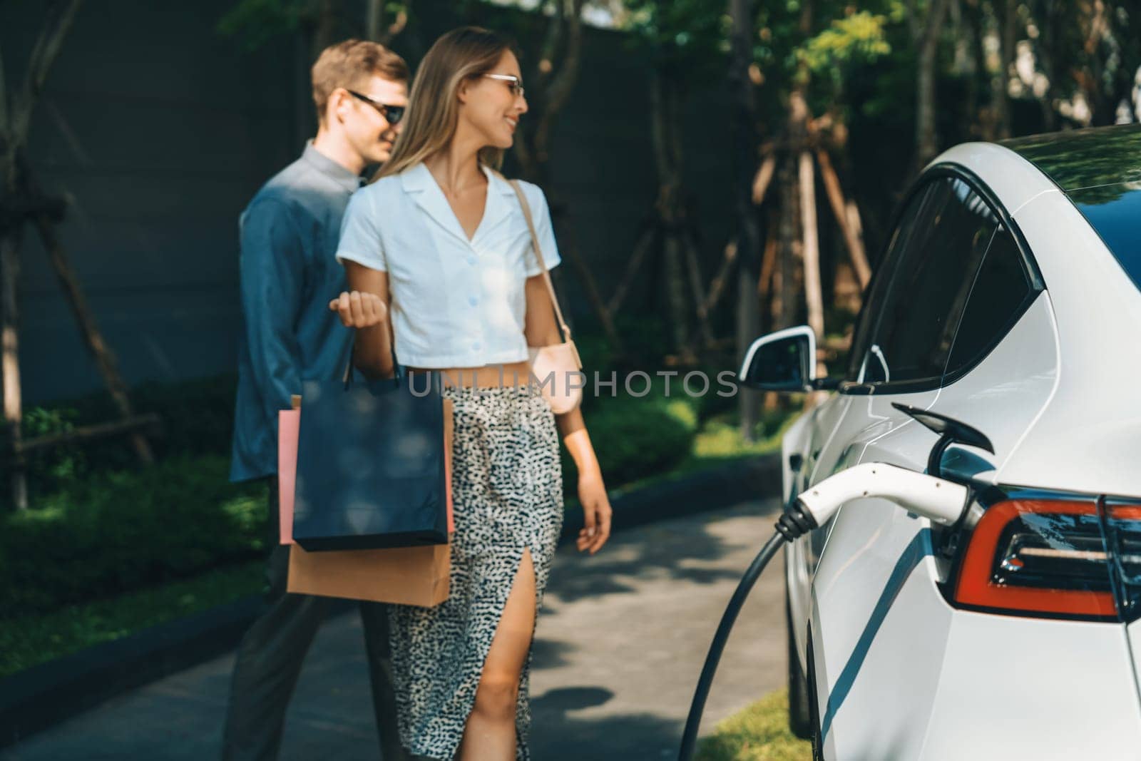 Young couple travel with EV electric car charging in green sustainable city outdoor garden in summer shows urban sustainability lifestyle by green clean rechargeable energy of electric vehicle innards