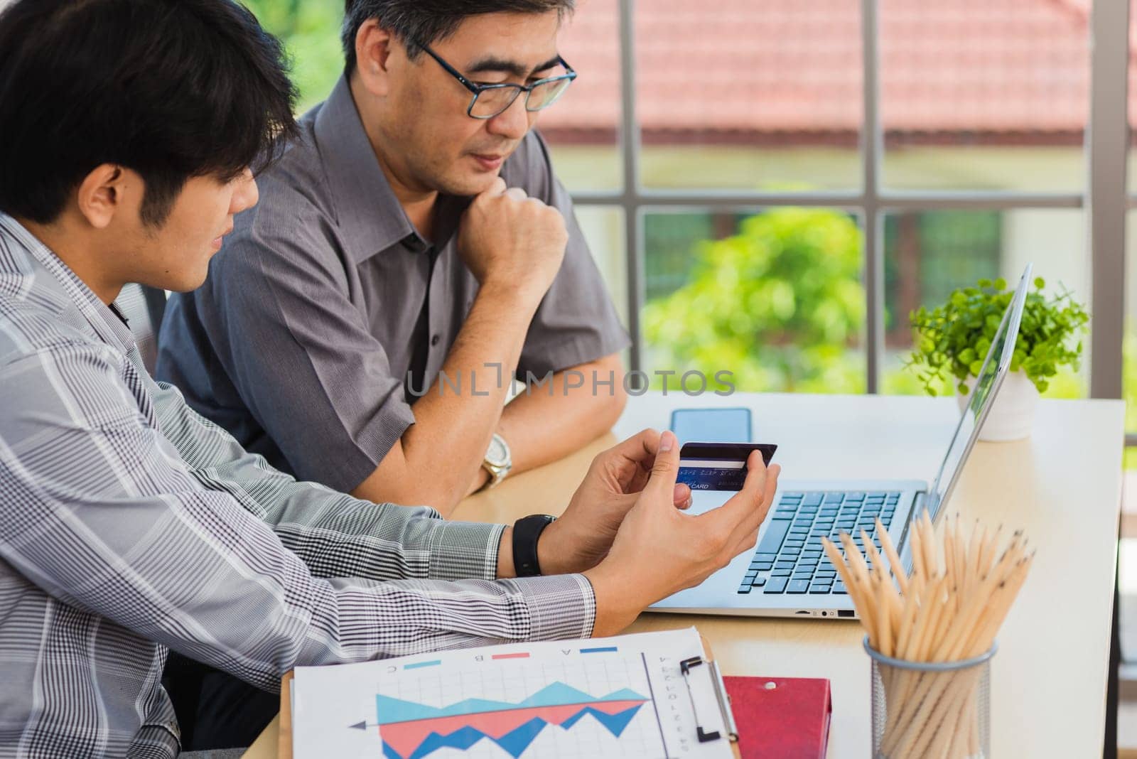 Asian young man teaching senior father how to use credit card for shopping online with laptop computer in the living room, adult son holding bank card helping Dad to purchases web online application