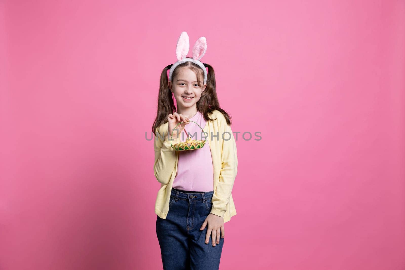 Charming little girl holding basket of eggs and other easter toys, pink background. Toddler expressing childhood innocence and the joyous anticipation of spring holiday festivities.
