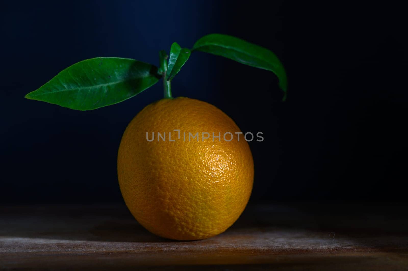 A bright orange sits elegantly on a wooden surface, its green leaf adding a touch of vitality against a dark background.