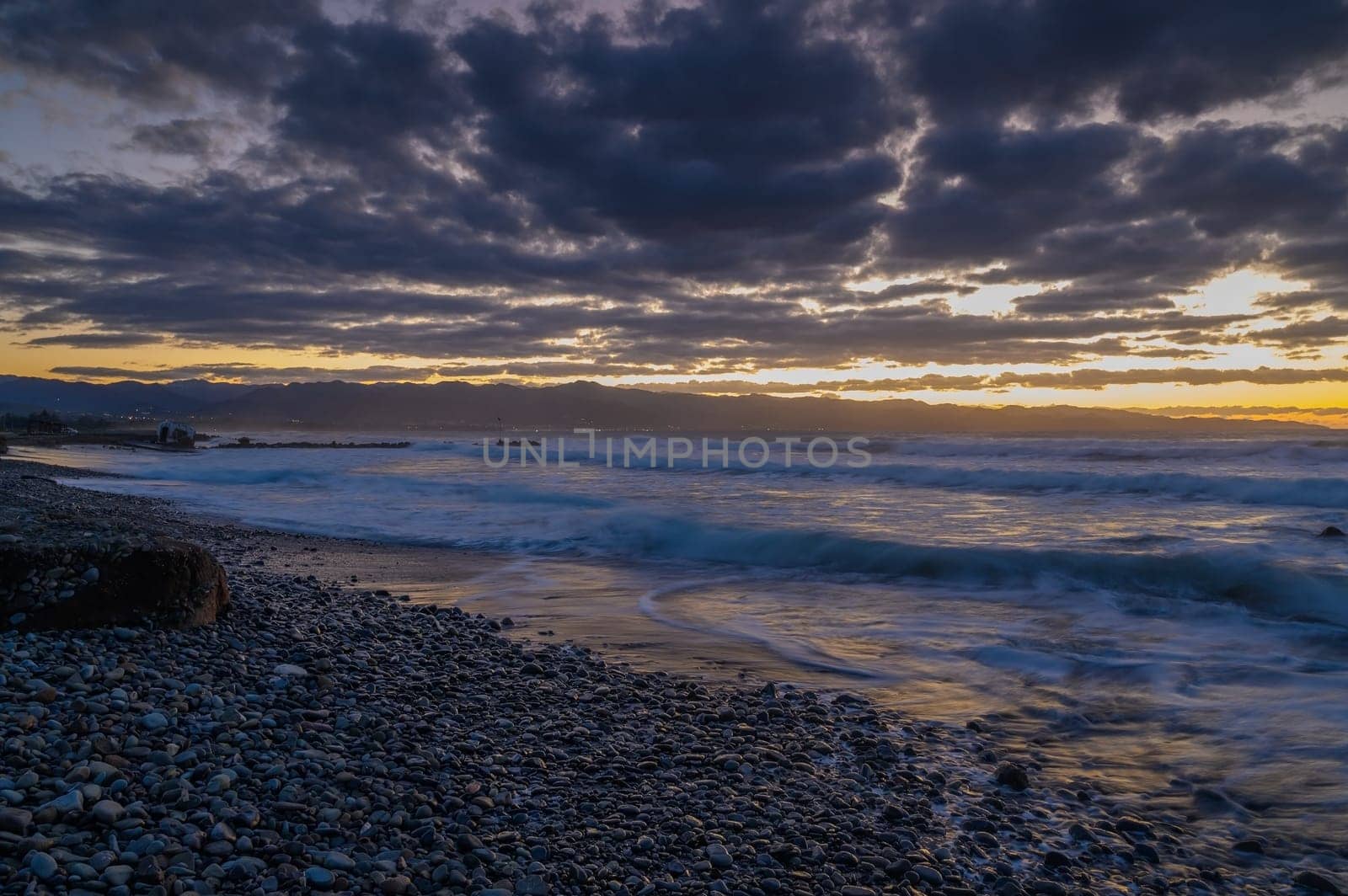 Golden hues reflect on water while waves gently roll over pebbles under a dramatic sky at sunset
