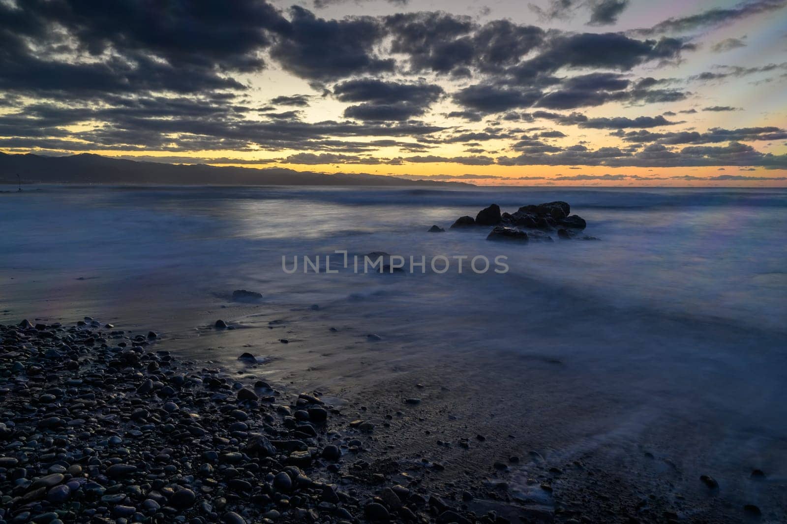 Waves softly lap at smooth stones along the shore as the sun sets, painting the sky with vibrant colors and clouds.