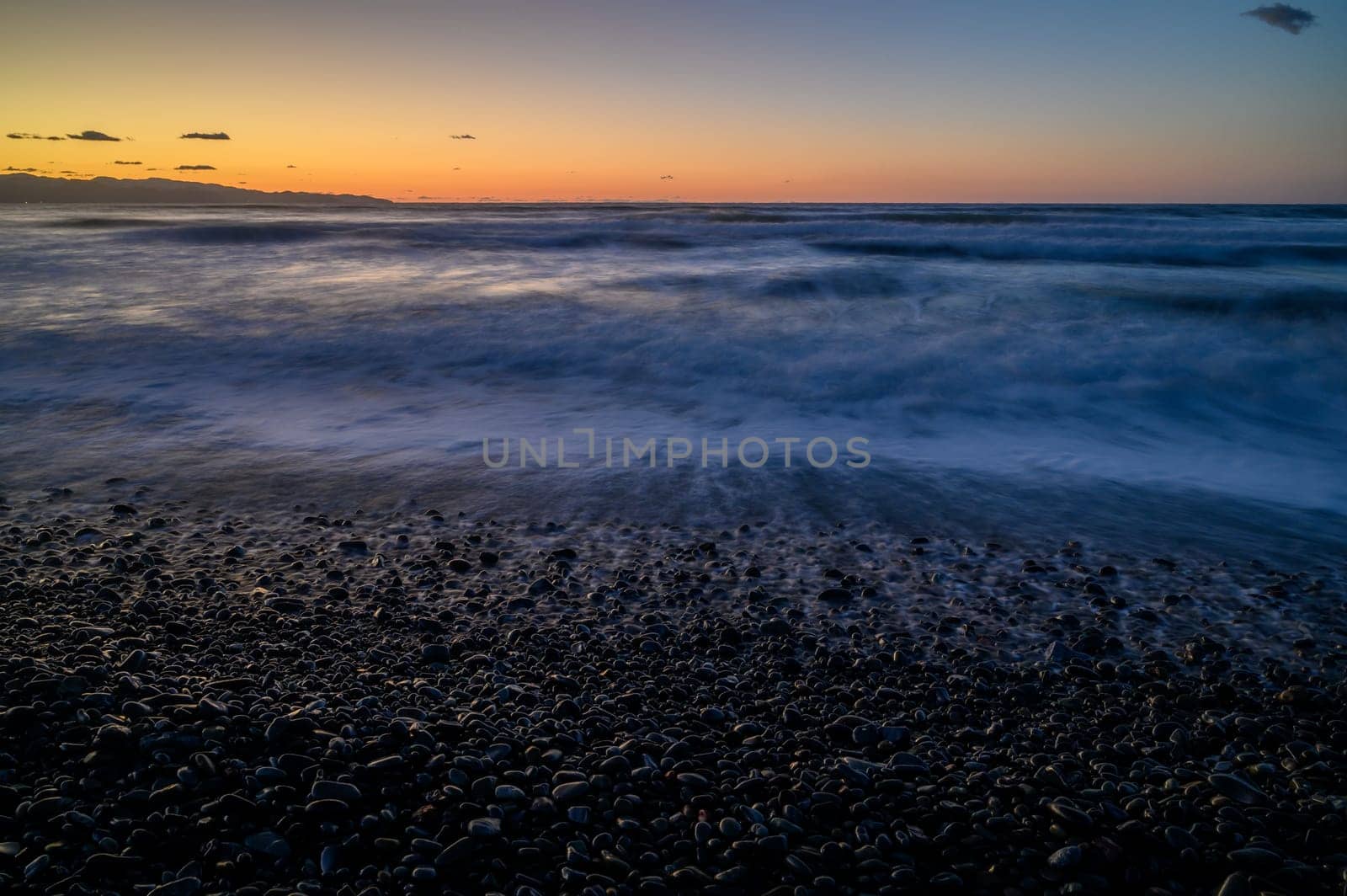 Gentle waves roll over smooth pebbles under a vivid twilight sky, creating a peaceful coastal atmosphere at sunset.