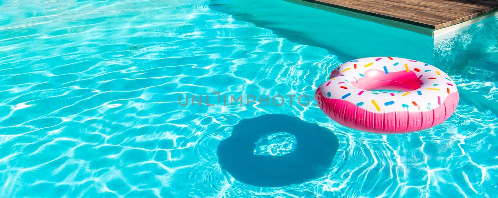 Pink donut inflatable float in turquoise swimming pool with shadow and wooden deck