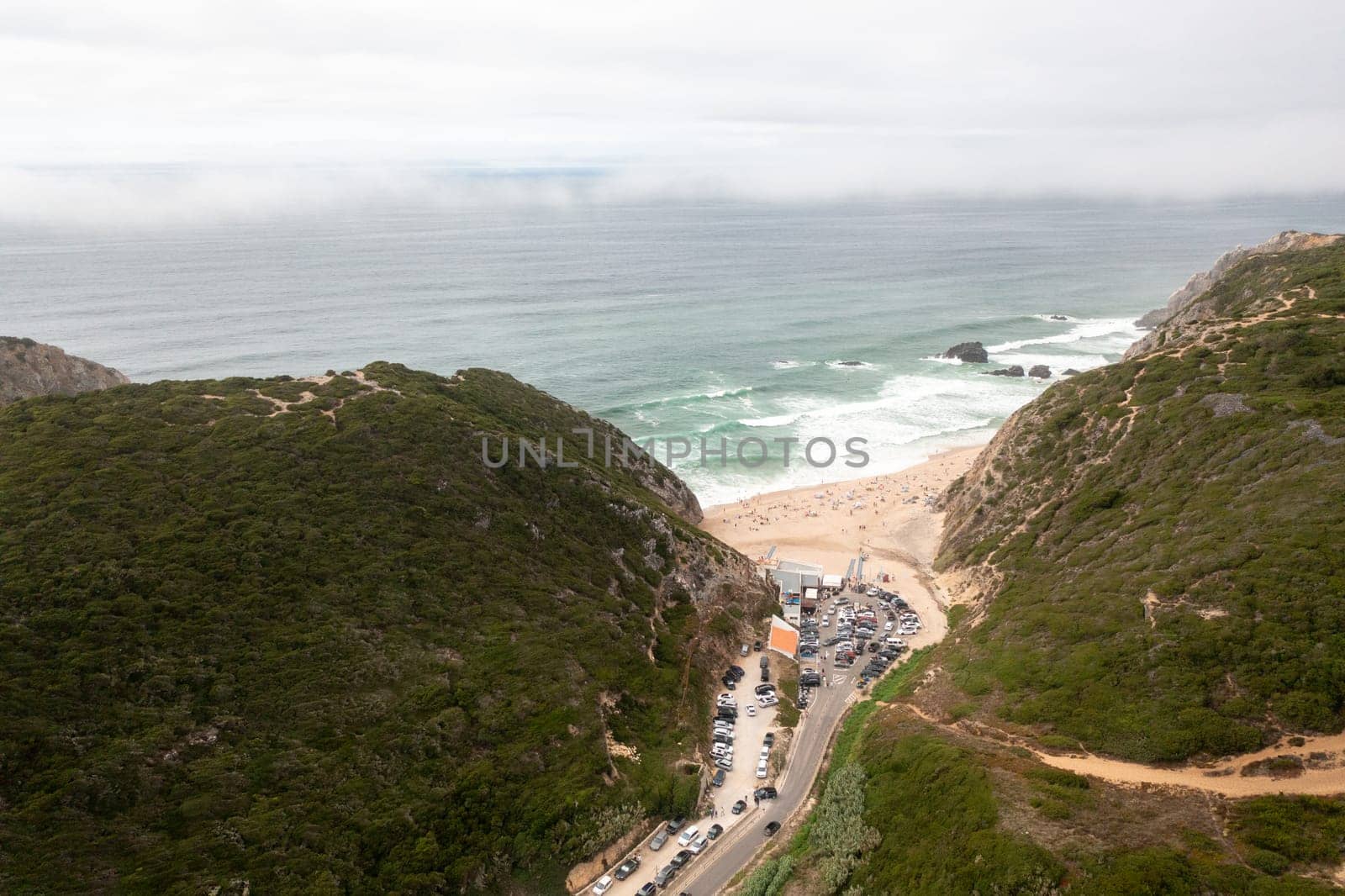 Aerial drone view of parking lot nestled between hills at Adraga Beach in Sintra, Portugal. The scenic landscape contrasts with orderly parked cars, highlighting natural beauty of coastal setting