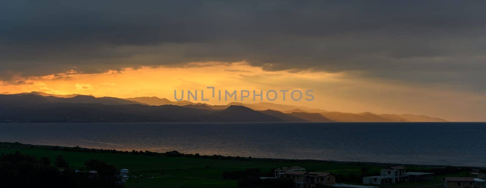 The sky glows with shades of gold as the sun sets behind rugged mountains by the tranquil sea.