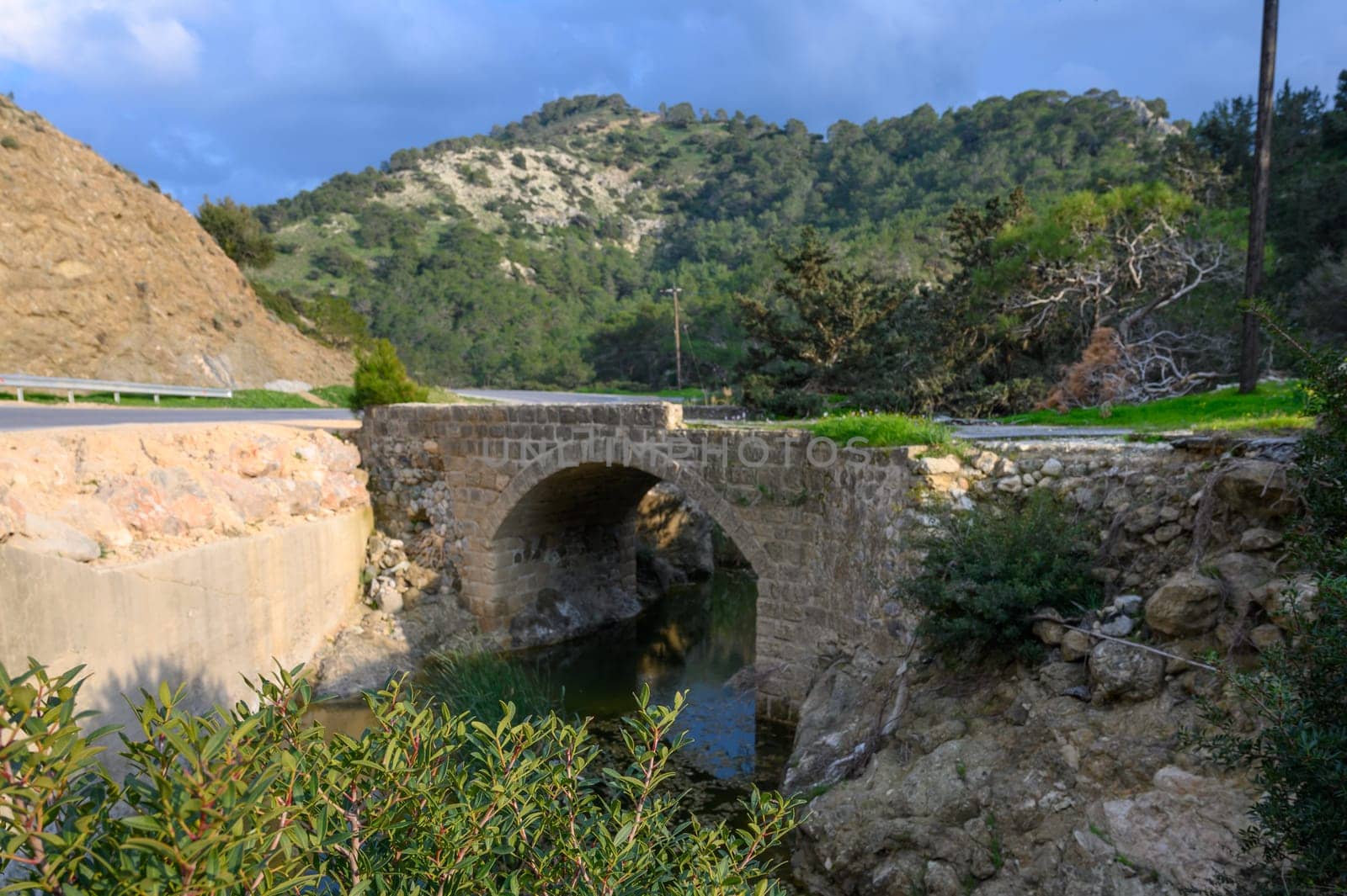 Sunlight filters through the clouds, casting a warm glow on a historic stone bridge arching over a tranquil river.