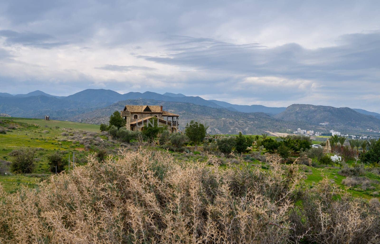 A charming stone house quietly rests amidst lush hills, framed by distant mountains and a dramatic sky at twilight.