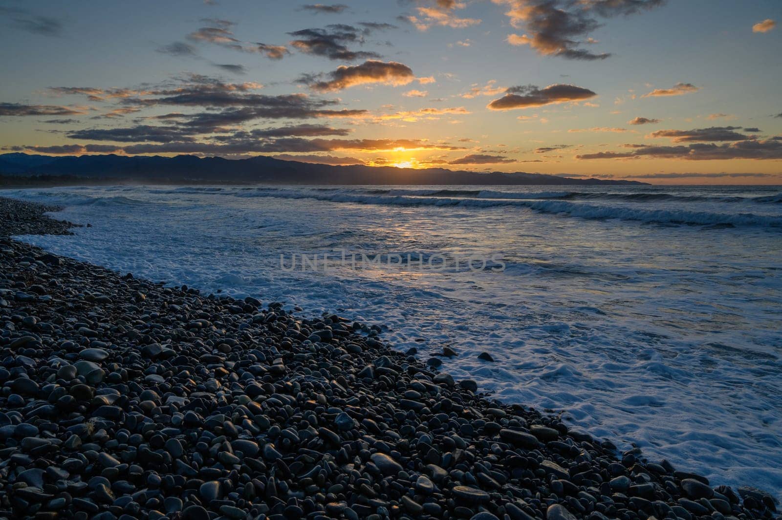 Golden sun descends on the horizon, illuminating pebbled beach while waves embrace the shore at twilight.