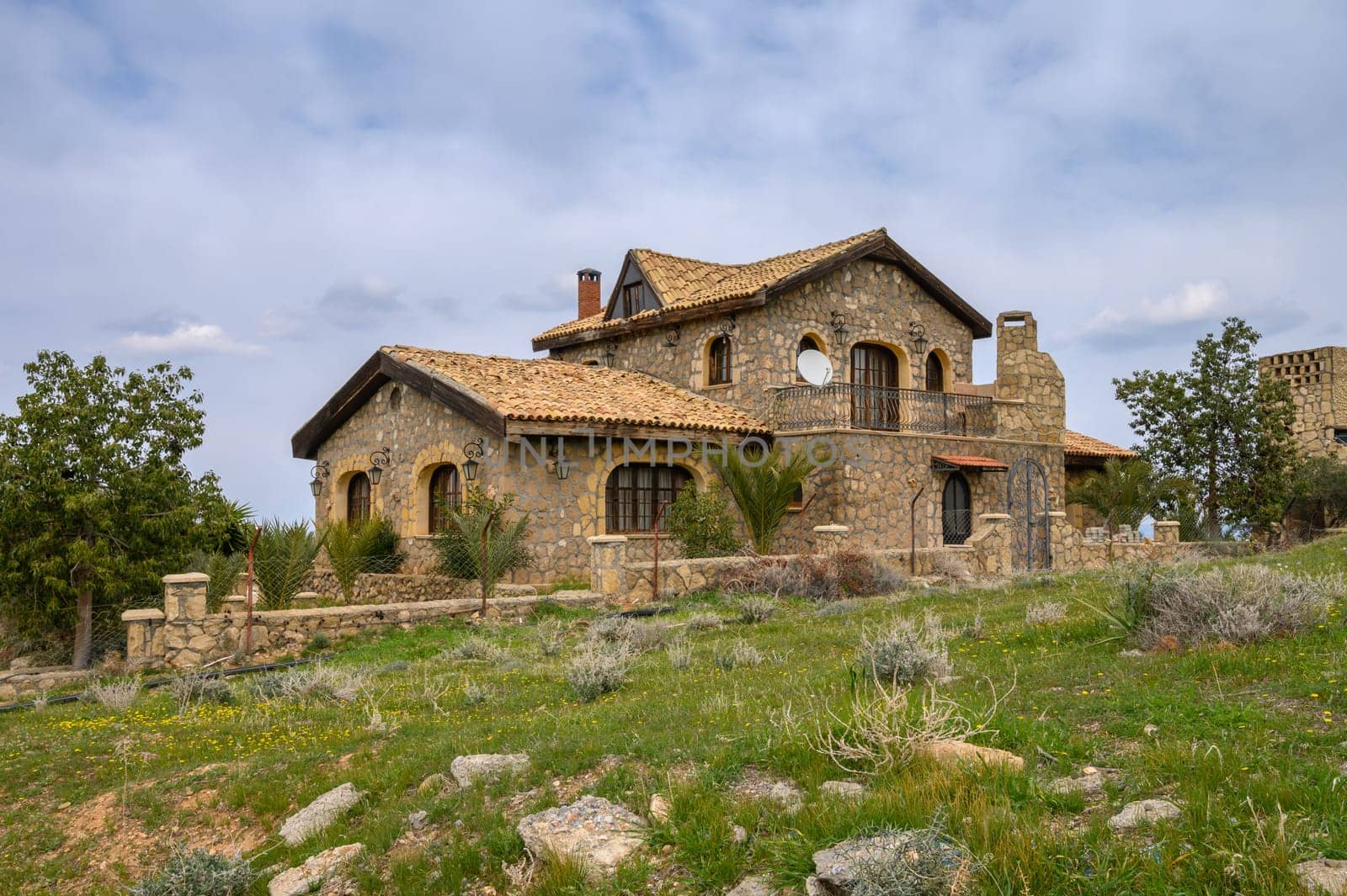 Charming villa with a rustic design sits amidst lush greenery under a cloudy sky, offering a peaceful escape.