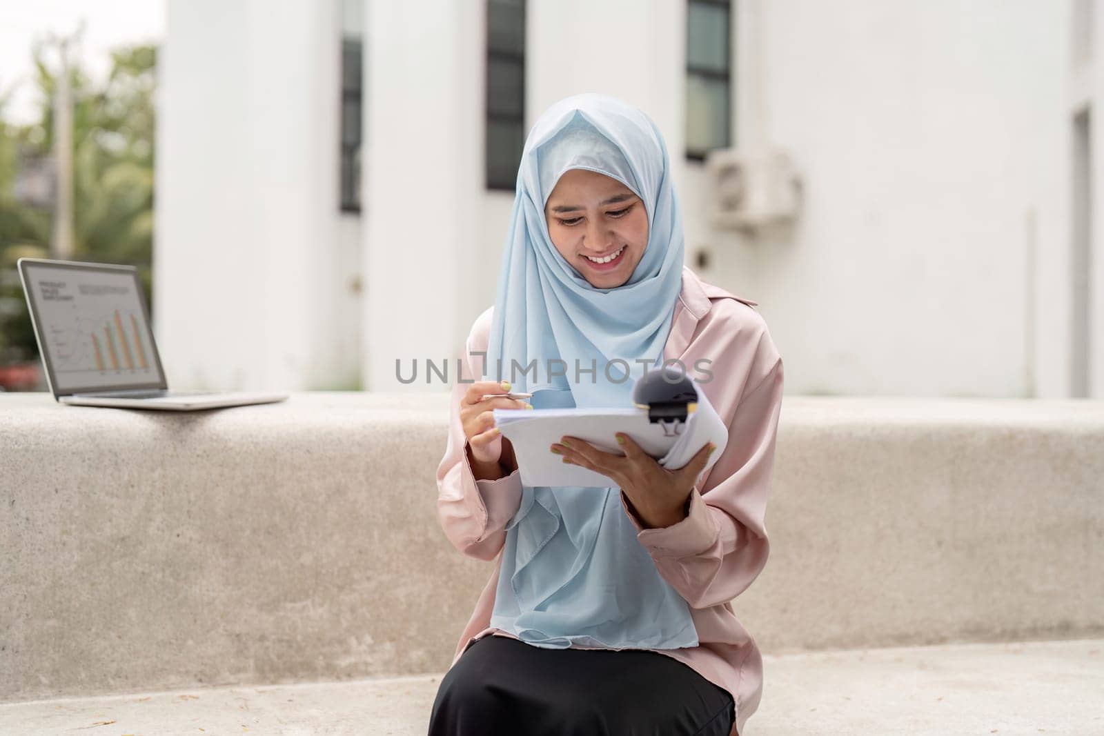 A joyful Muslim businesswoman reviews data on her tablet, sitting beside a laptop, showcasing dedication to her work and cultural diversity in a contemporary setting.