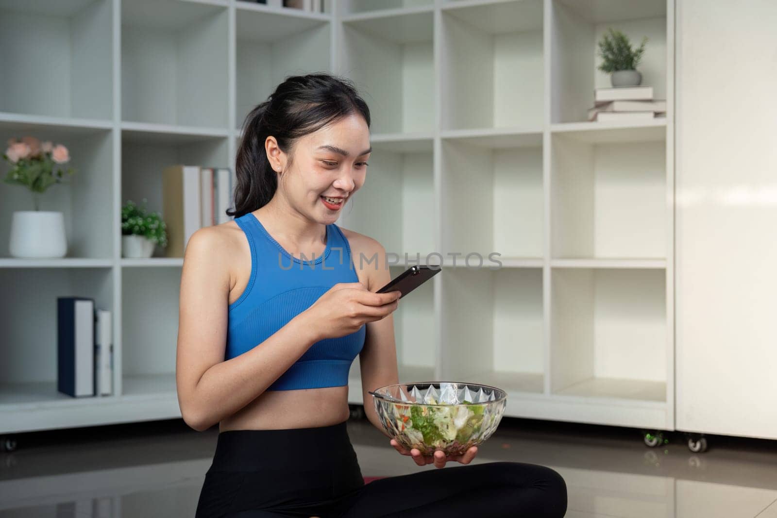 A woman enjoying a colorful salad and using her smartphone while sitting in a stylish living area, blending healthy eating with technology.