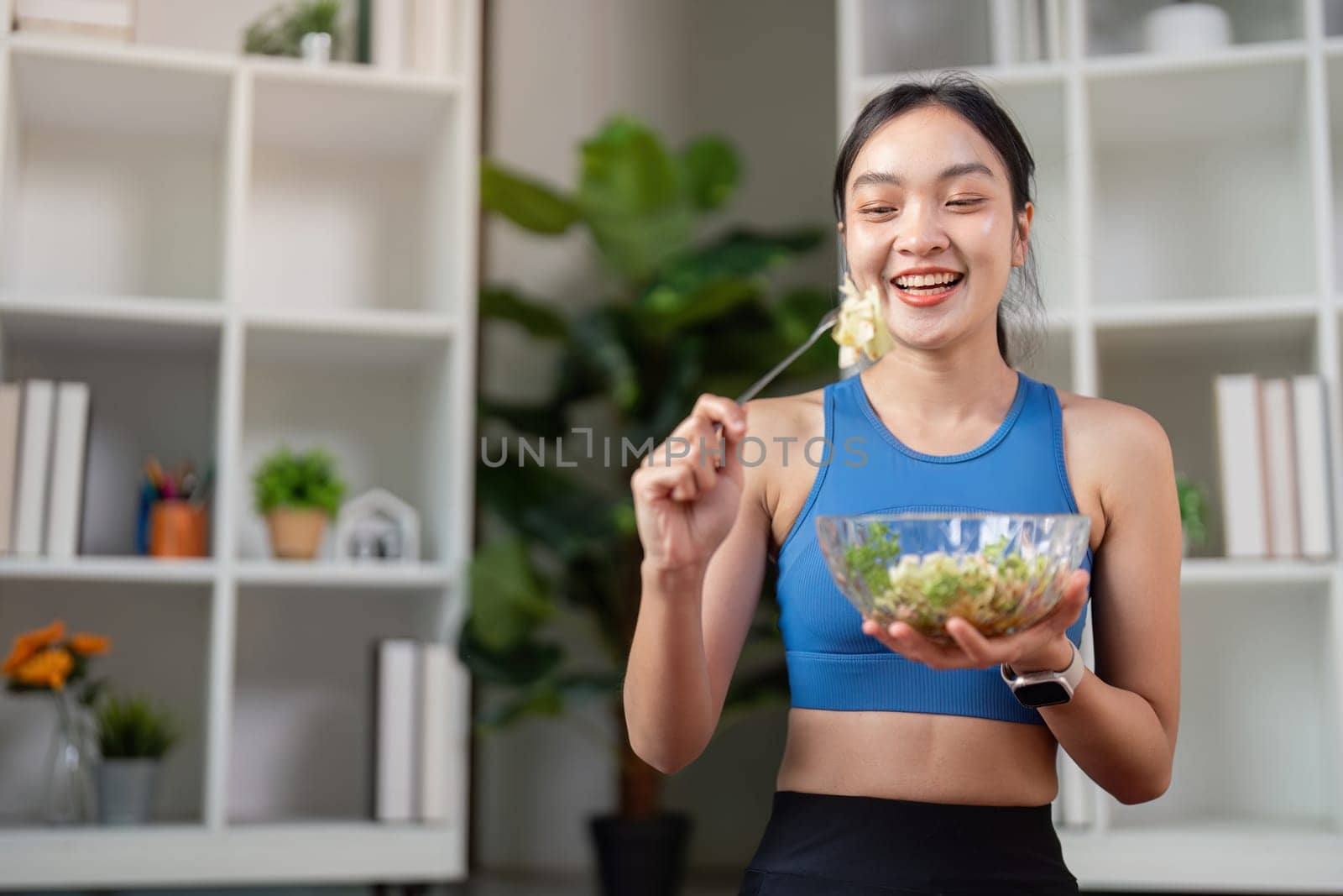 A cheerful woman in athletic clothing happily prepares to eat a nutritious salad, promoting healthy living in a stylish home environment.